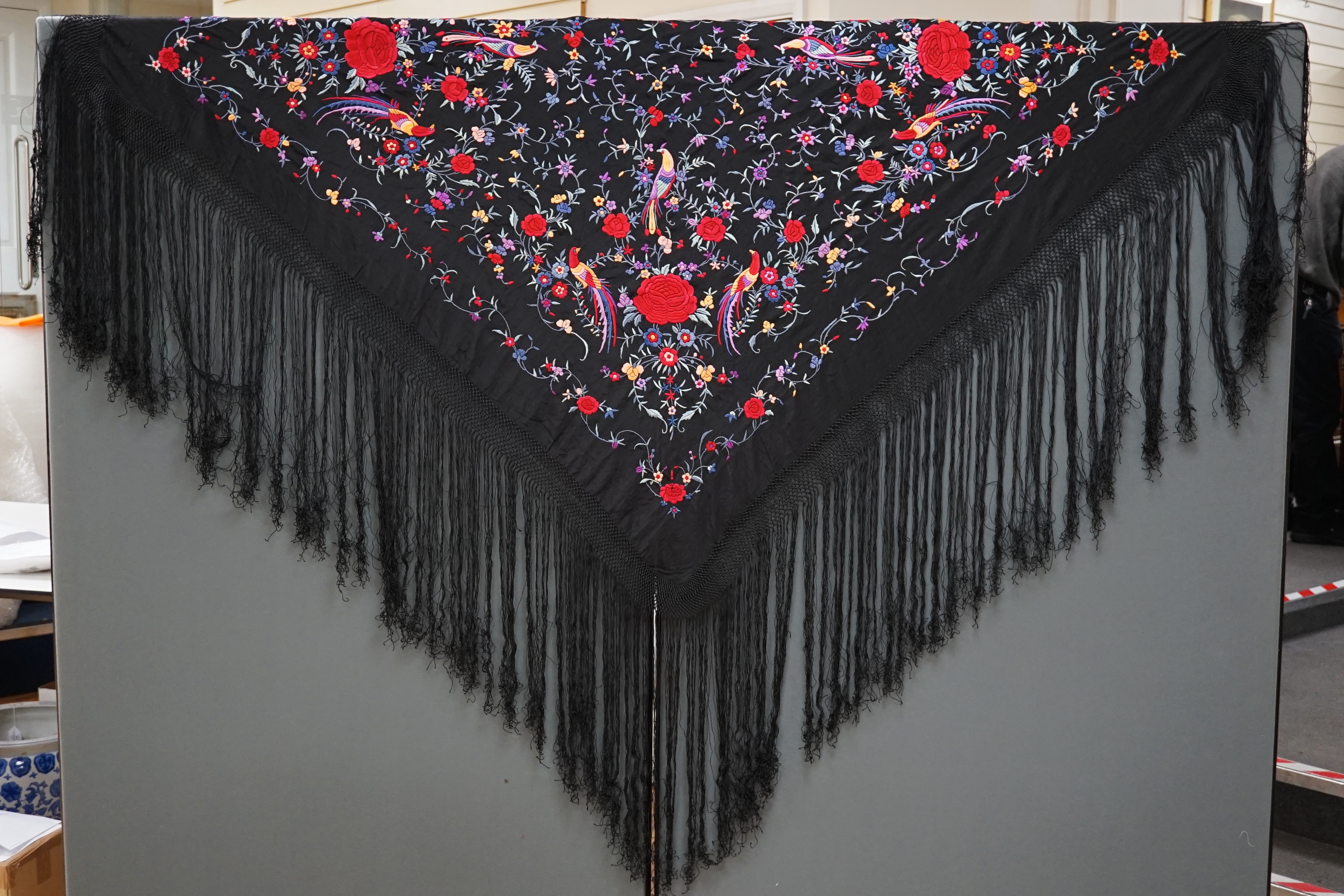 An early 20th century Chinese black silk fringed shawl with polychrome silk embroidery, embroidered with meandering floral design, the corners embroidered with three brightly coloured birds of paradise, large red roses a